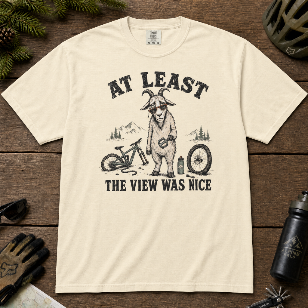 Ivory white t-shirt of a goat holding a bicycle pedal with a broken mountain bike surrounding it. Included text, "At Least the View Was Nice".