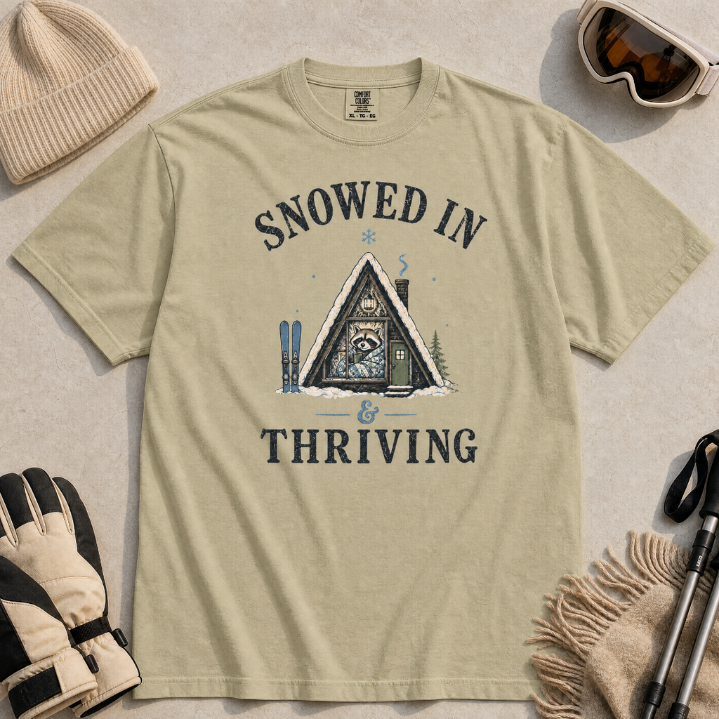 Flat lay of a bay green t-shirt featuring a retro “Snowed In & Thriving” raccoon cabin graphic, styled with winter accessories like ski goggles, gloves, and poles.