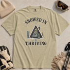Flat lay of a bay green t-shirt featuring a retro “Snowed In & Thriving” raccoon cabin graphic, styled with winter accessories like ski goggles, gloves, and poles.