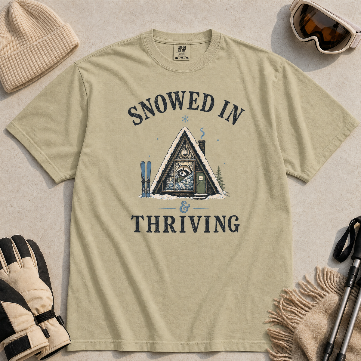 Flat lay of a bay green t-shirt featuring a retro “Snowed In & Thriving” raccoon cabin graphic, styled with winter accessories like ski goggles, gloves, and poles.