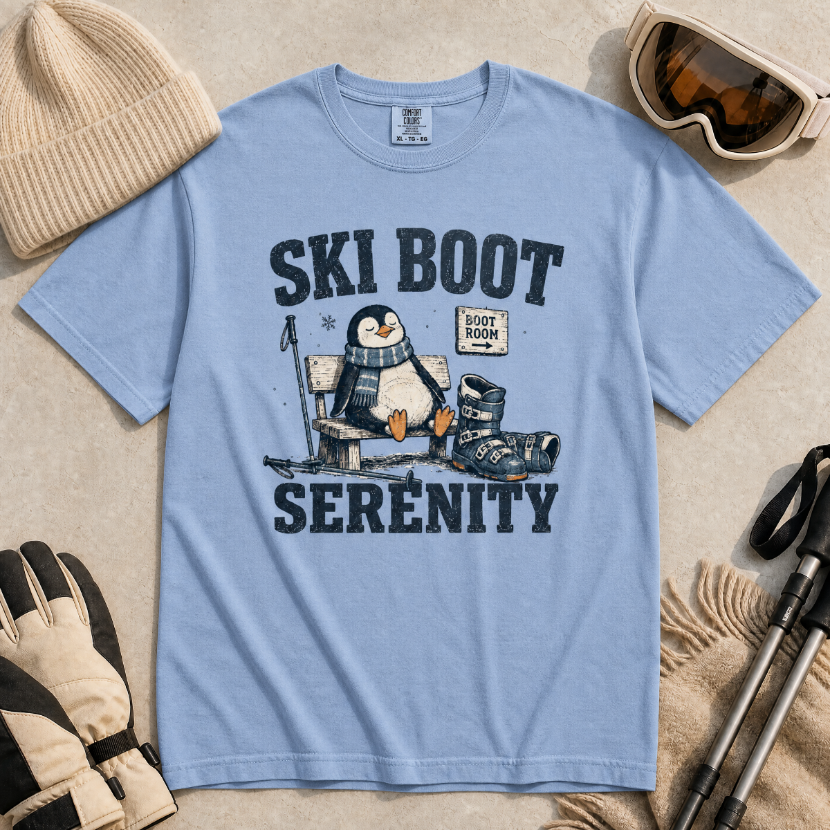 Flat lay of a washed denim blue t-shirt featuring a retro “Ski Boot Serenity” penguin design, styled with winter accessories like ski goggles, gloves, and poles.