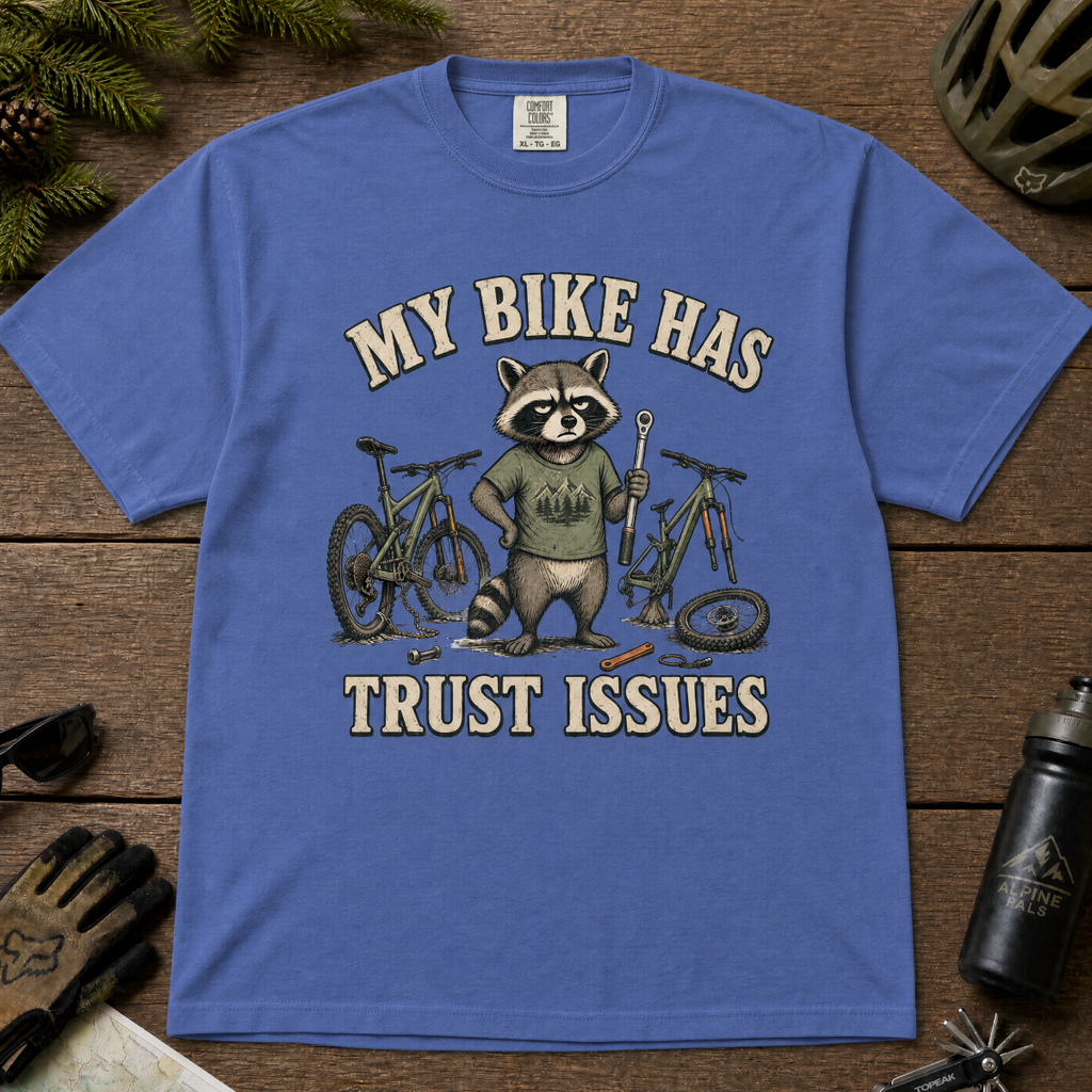 Mystic blue t-shirt with a raccoon graphic and text 'My bike has trust issues' on a wooden surface with cycling accessories.
