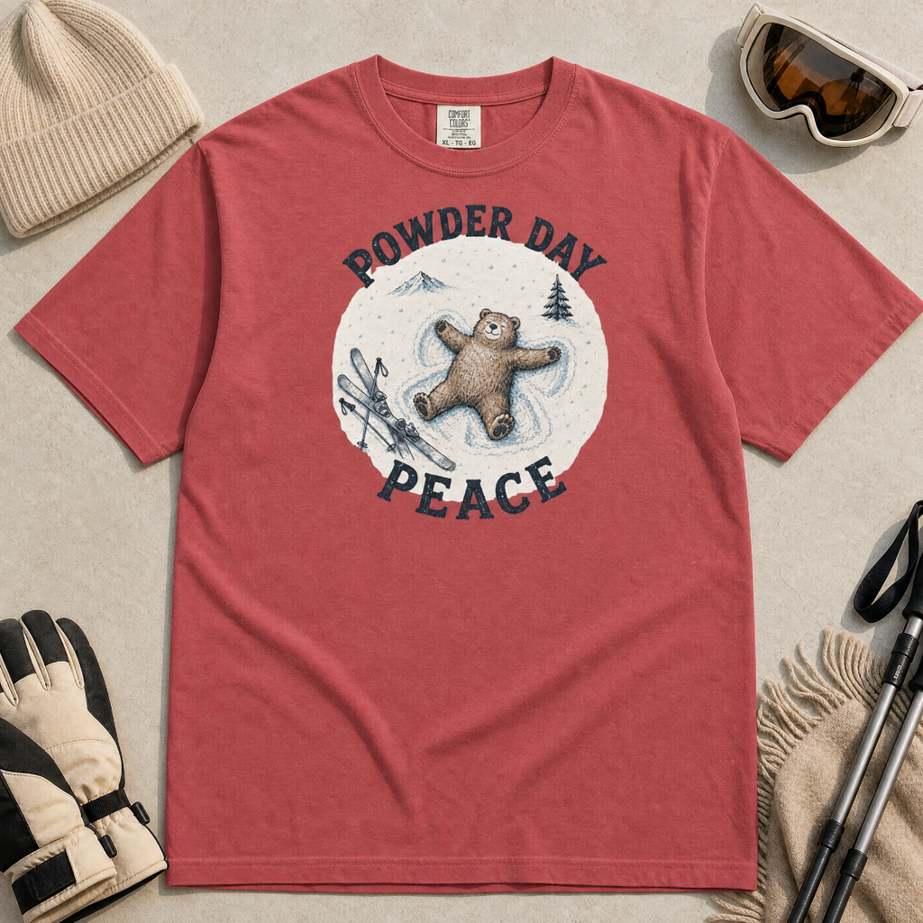 Flat lay of a crimson red t-shirt featuring a peaceful bear snow angel ski graphic with “Powder Day Peace” text, styled with winter accessories.