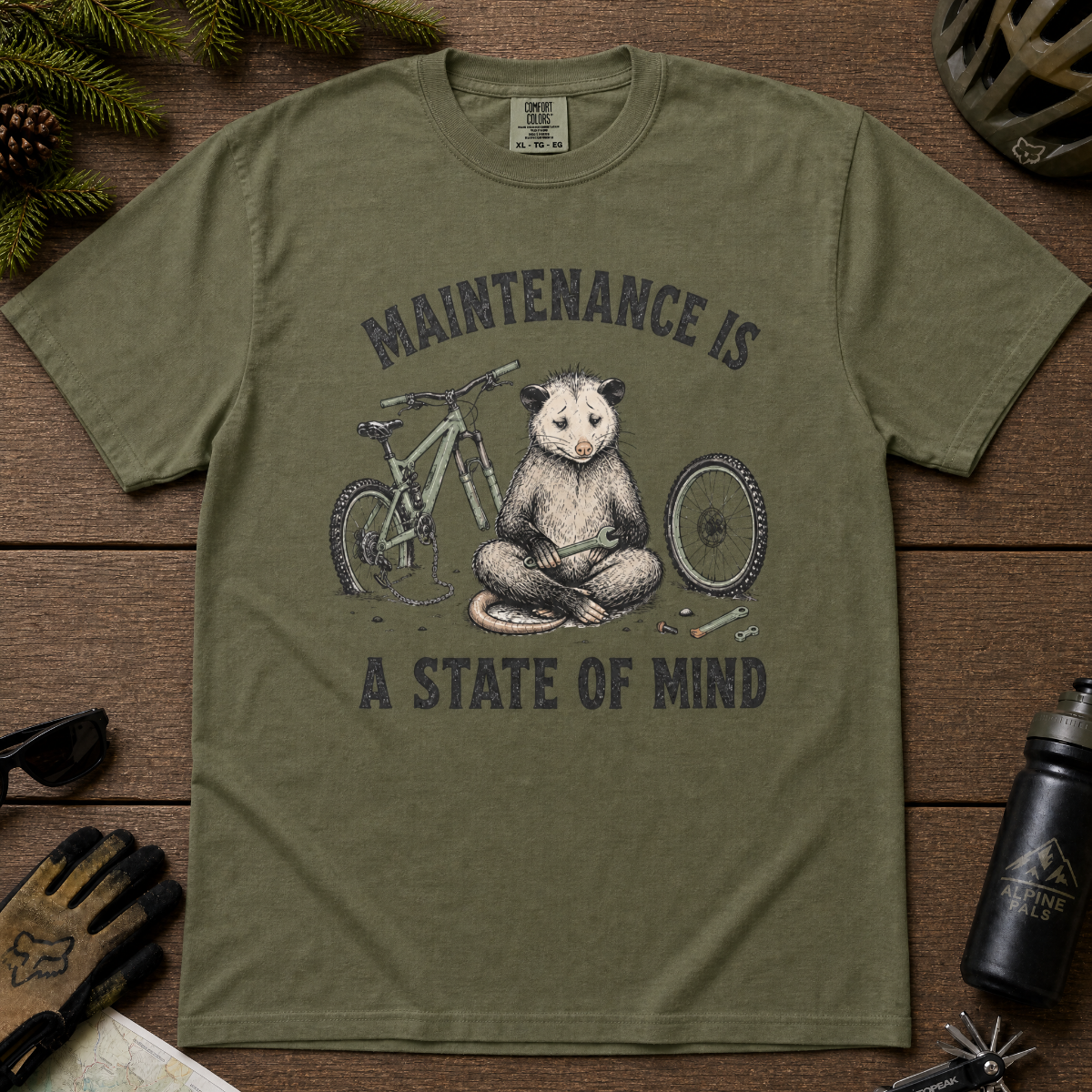 Hemp green t-shirt featuring a possum meditating while fixing a bike with text, "Maintenance is a State of Mind".