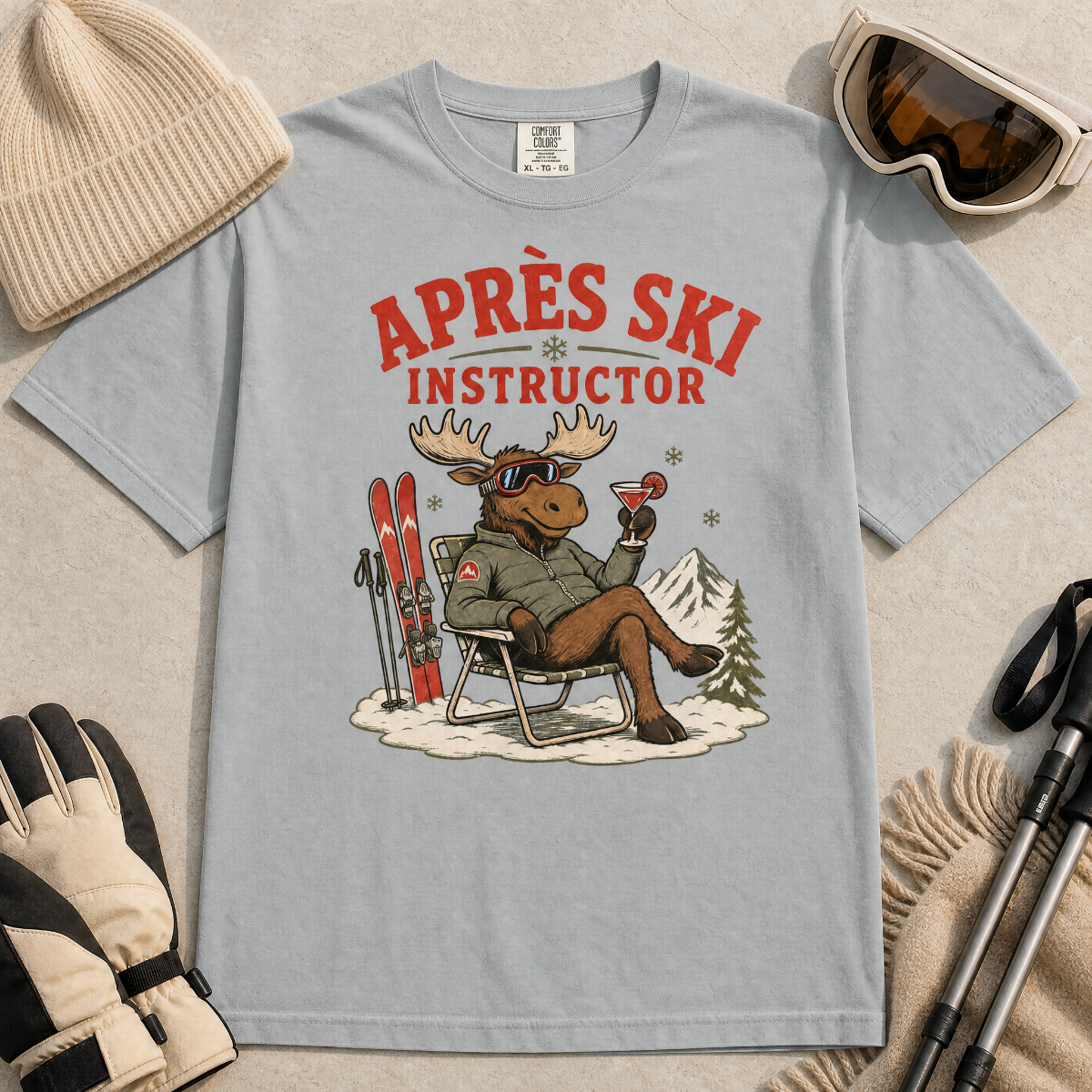 Granite gray t-shirt with a moose graphic and 'Après Ski Instructor' text, surrounded by skiing accessories on a beige background.