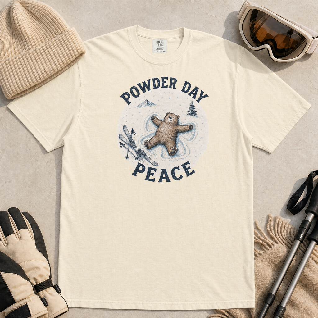 Flat lay of an ivory white t-shirt featuring a peaceful bear snow angel ski graphic with “Powder Day Peace” text, styled with winter accessories.