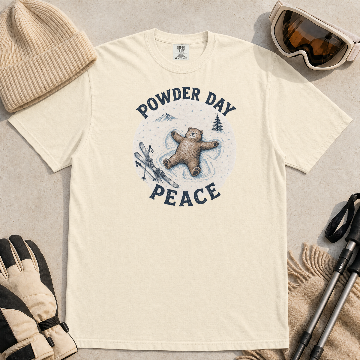 Flat lay of an ivory white t-shirt featuring a peaceful bear snow angel ski graphic with “Powder Day Peace” text, styled with winter accessories.