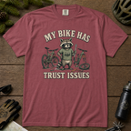 Brick red t-shirt with raccoon graphic and 'My bike has trust issues' text on a wooden surface with cycling accessories.