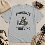 Flat lay of a granite gray t-shirt featuring a retro “Snowed In & Thriving” raccoon cabin graphic, styled with winter accessories like ski goggles, gloves, and poles.