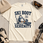Flat lay of an ivory white t-shirt featuring a retro “Ski Boot Serenity” penguin design, styled with winter accessories like ski goggles, gloves, and poles.