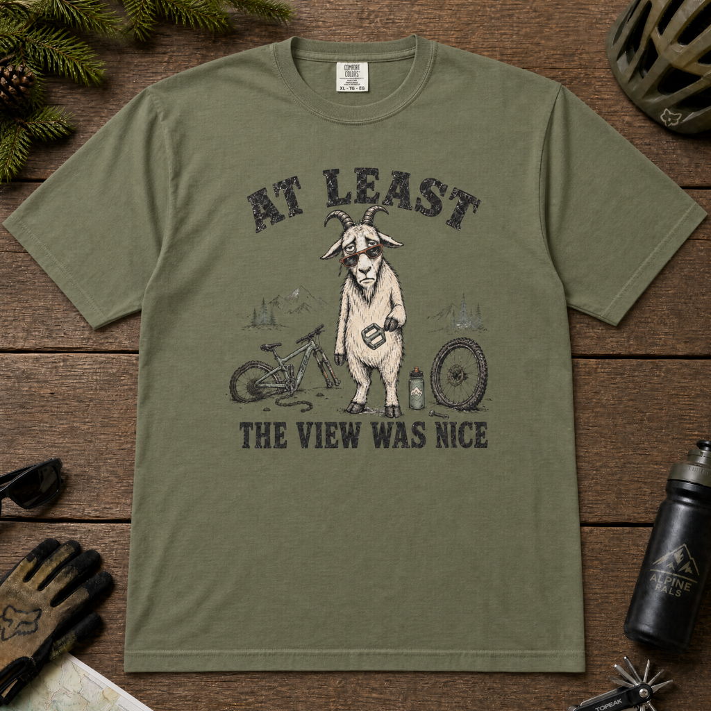 Moss green t-shirt of a goat holding a bicycle pedal with a broken mountain bike surrounding it. Included text, "At Least the View Was Nice".