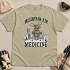 Flat lay of a khaki colored t-shirt featuring a peaceful moose ski graphic with “Mountain Air Medicine” text, styled with winter accessories.