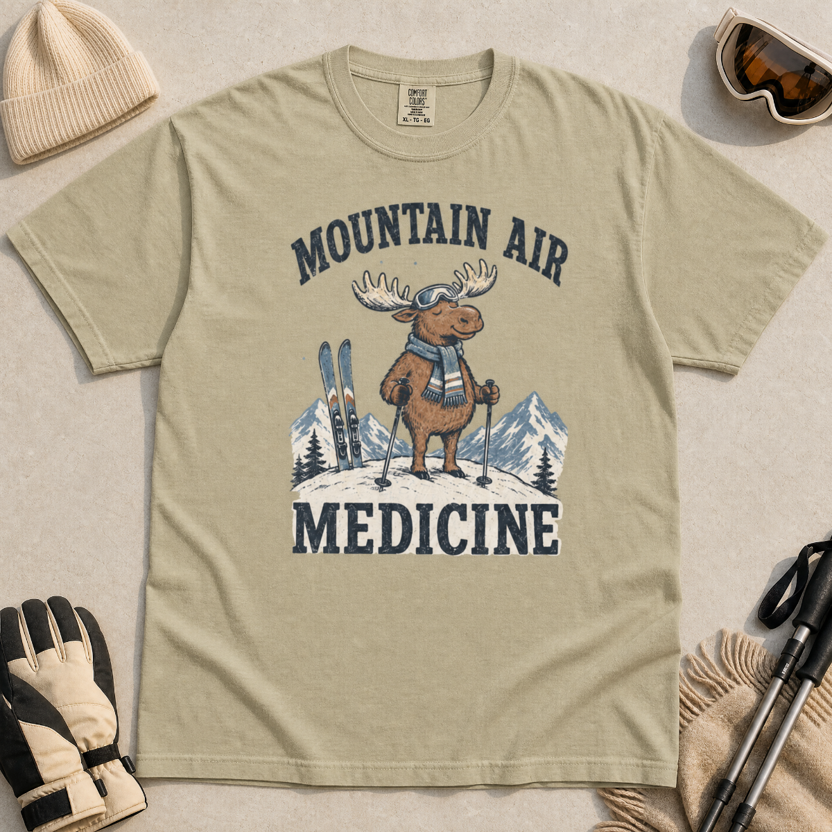 Flat lay of a khaki colored t-shirt featuring a peaceful moose ski graphic with “Mountain Air Medicine” text, styled with winter accessories.