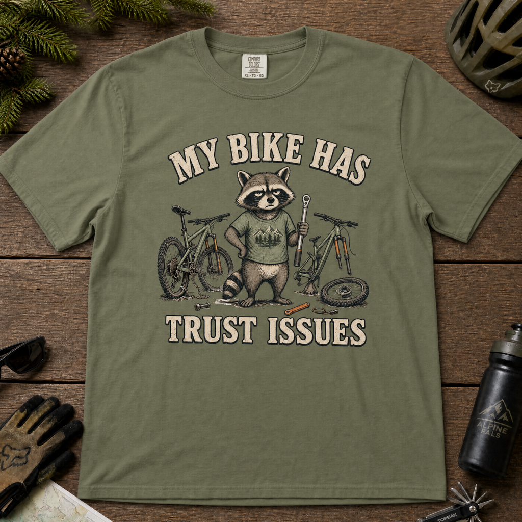 Moss green t-shirt with a raccoon graphic and text 'My bike has trust issues' on a wooden surface with cycling accessories.