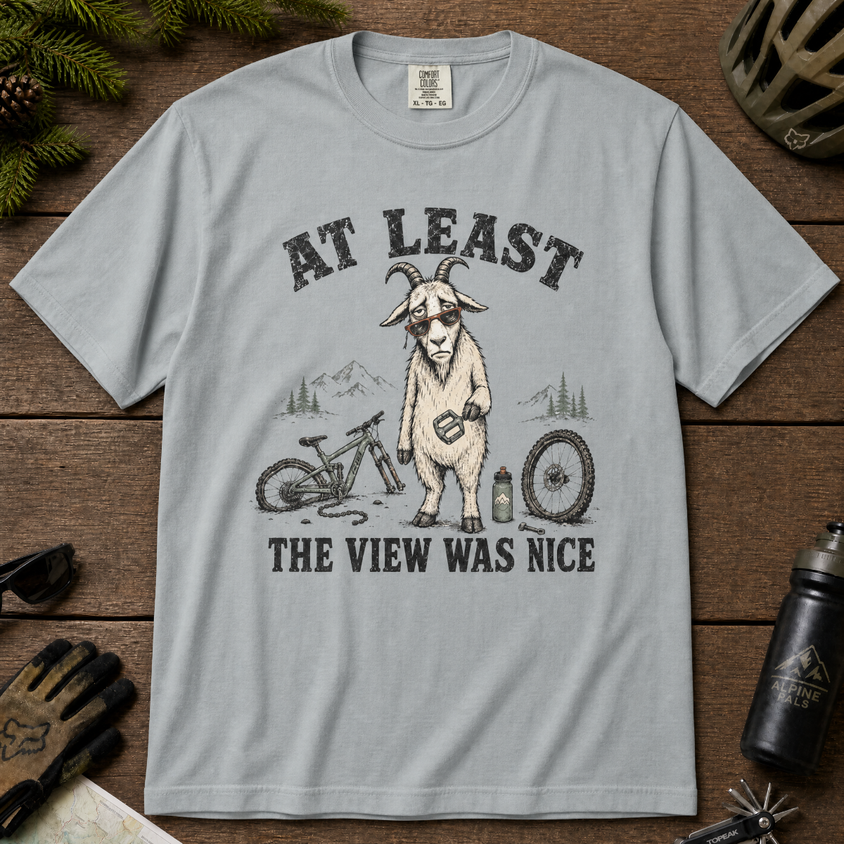 Granite gray t-shirt of a goat holding a bicycle pedal with a broken mountain bike surrounding it. Included text, "At Least the View Was Nice".