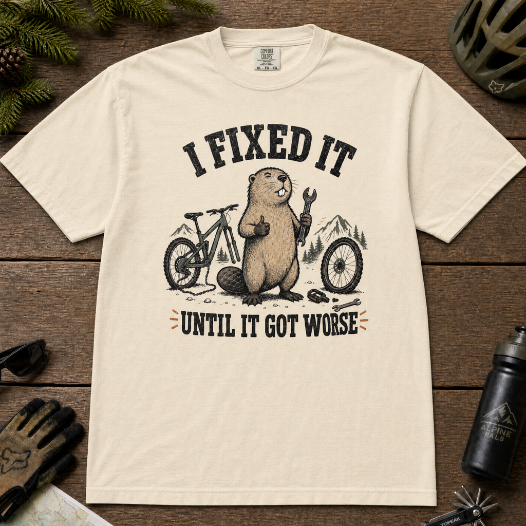 Ivory white t-shirt with graphic of beaver and text 'I Fixed It Until It Got Worse' on a wooden surface with cycling accessories.
