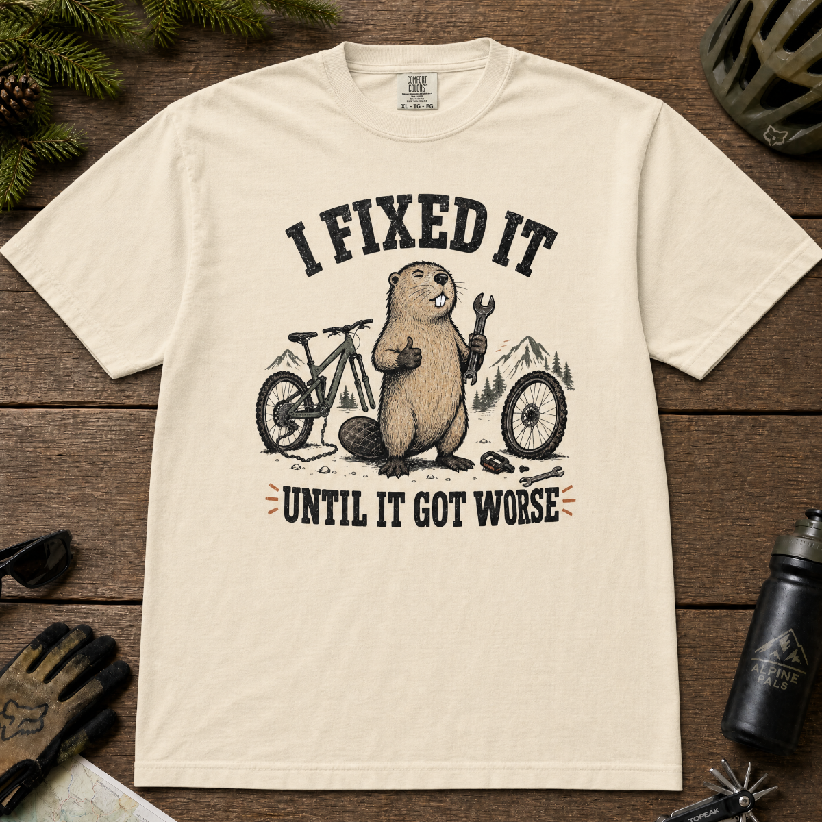 Ivory white t-shirt with graphic of beaver and text 'I Fixed It Until It Got Worse' on a wooden surface with cycling accessories.
