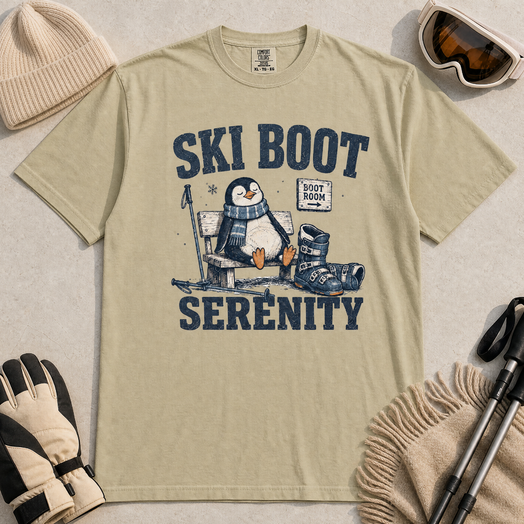 Flat lay of a khaki colored t-shirt featuring a retro “Ski Boot Serenity” penguin design, styled with winter accessories like ski goggles, gloves, and poles.