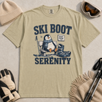 Flat lay of a khaki colored t-shirt featuring a retro “Ski Boot Serenity” penguin design, styled with winter accessories like ski goggles, gloves, and poles.