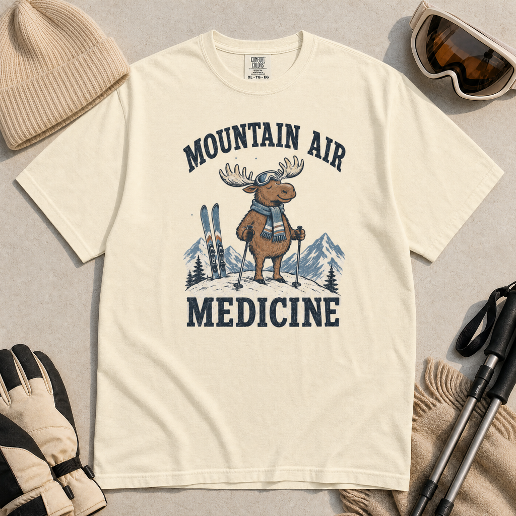 Flat lay of a ivory white t-shirt featuring a peaceful moose ski graphic with “Mountain Air Medicine” text, styled with winter accessories.