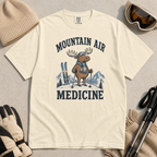 Flat lay of a ivory white t-shirt featuring a peaceful moose ski graphic with “Mountain Air Medicine” text, styled with winter accessories.