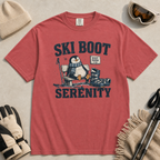 Flat lay of a crimson red t-shirt featuring a retro “Ski Boot Serenity” penguin design, styled with winter accessories like ski goggles, gloves, and poles.