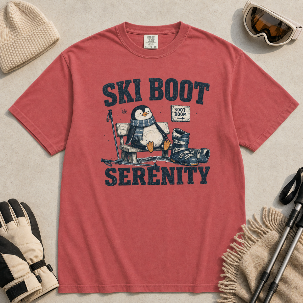 Flat lay of a crimson red t-shirt featuring a retro “Ski Boot Serenity” penguin design, styled with winter accessories like ski goggles, gloves, and poles.