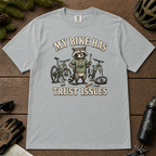 Granite gray t-shirt with a raccoon graphic and text 'My bike has trust issues' on a wooden surface with cycling accessories.