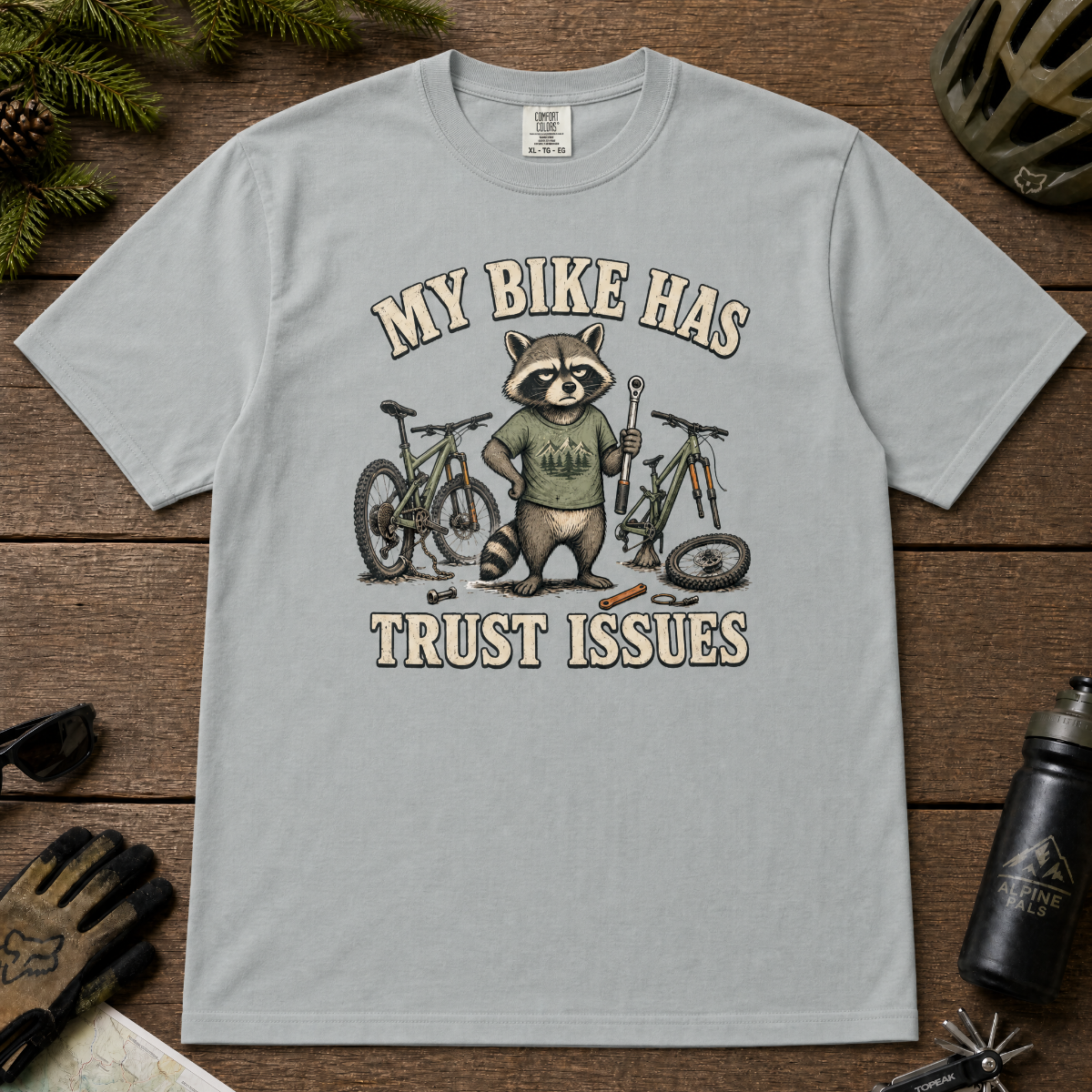 Granite gray t-shirt with a raccoon graphic and text 'My bike has trust issues' on a wooden surface with cycling accessories.