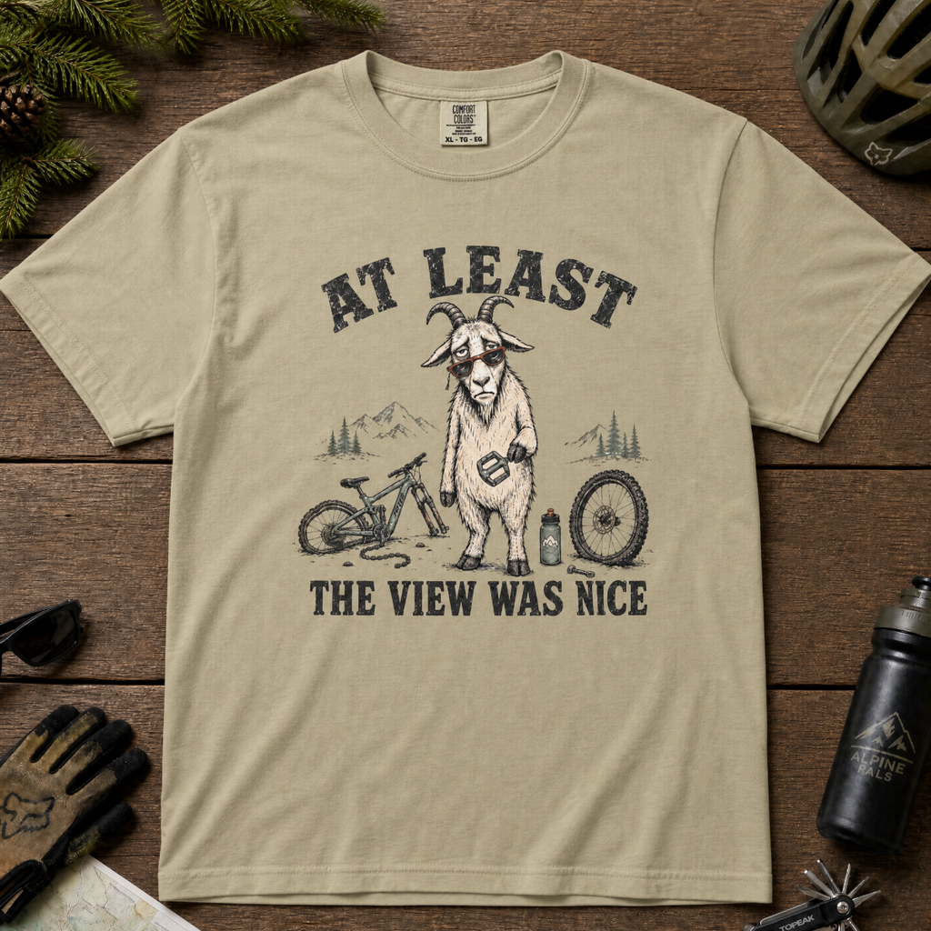 Khaki colored t-shirt of a goat holding a bicycle pedal with a broken mountain bike surrounding it. Included text, "At Least the View Was Nice".