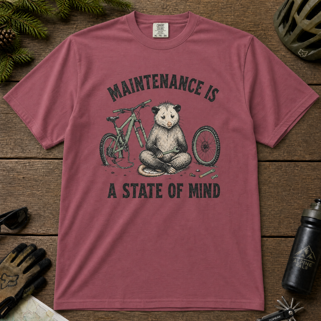 Brick red t-shirt featuring a possum meditating while fixing a bike with text, "Maintenance is a State of Mind".