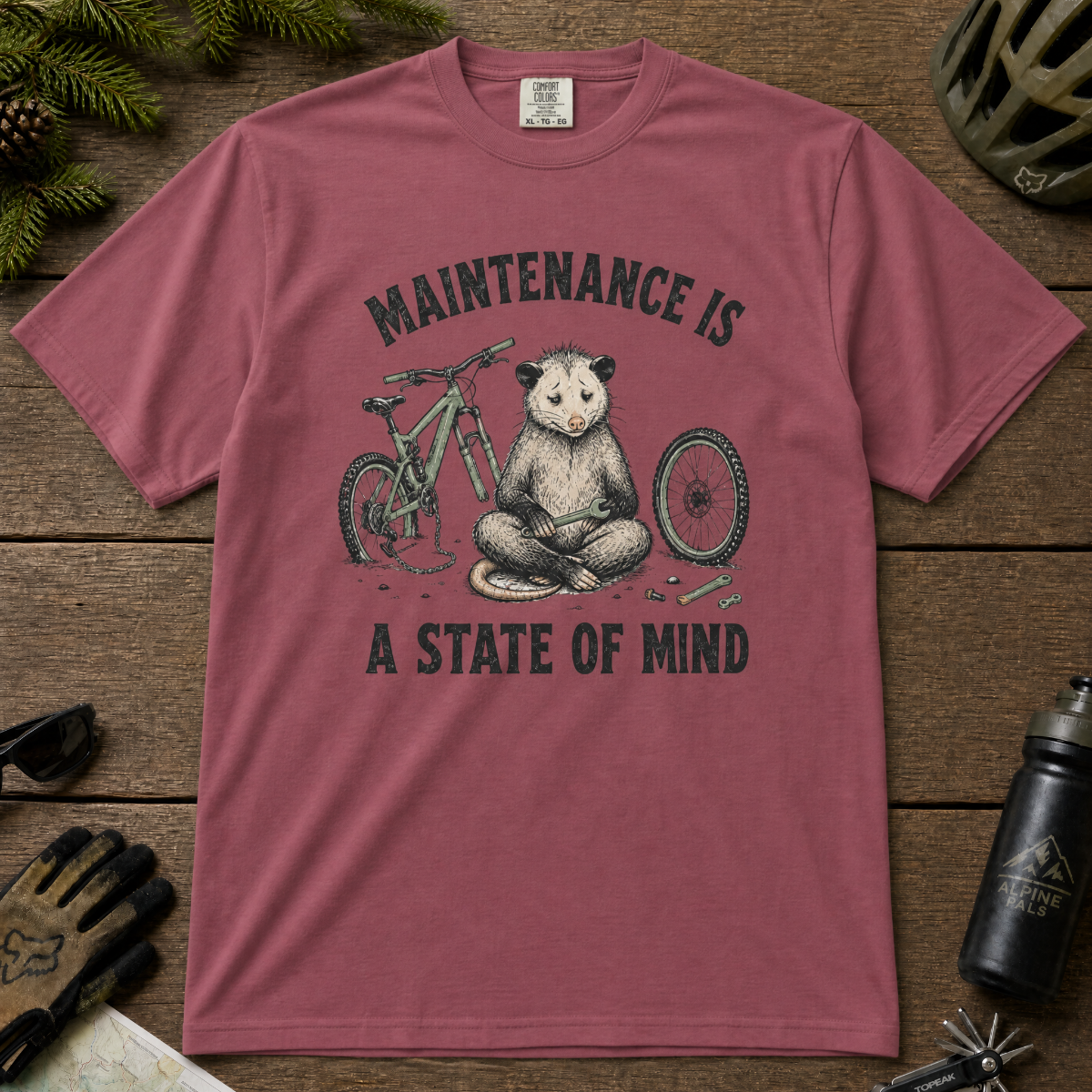 Brick red t-shirt featuring a possum meditating while fixing a bike with text, "Maintenance is a State of Mind".