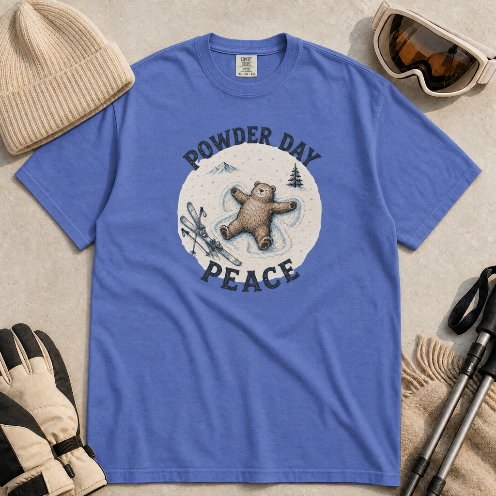 Flat lay of a mystic blue t-shirt featuring a peaceful bear snow angel ski graphic with “Powder Day Peace” text, styled with winter accessories.