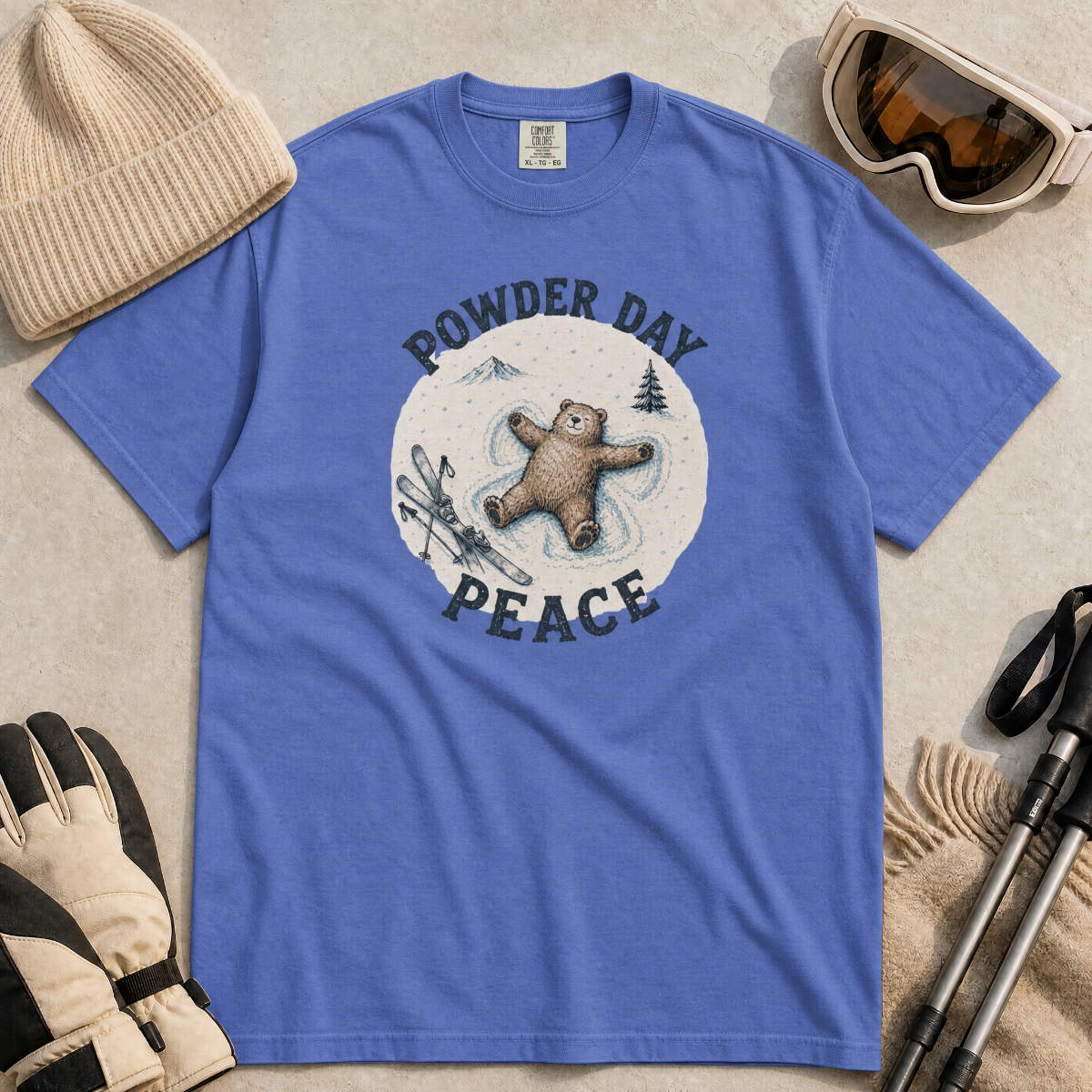 Flat lay of a mystic blue t-shirt featuring a peaceful bear snow angel ski graphic with “Powder Day Peace” text, styled with winter accessories.
