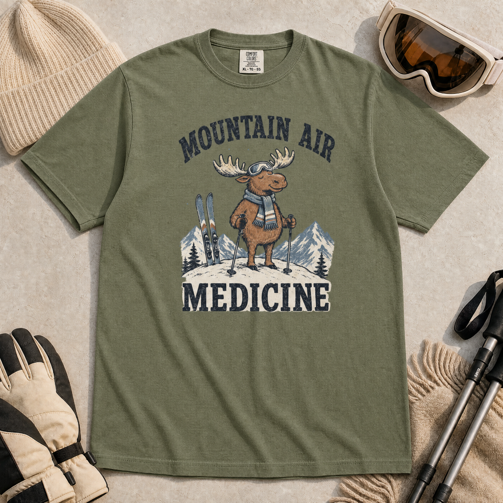 Flat lay of a moss green t-shirt featuring a peaceful moose ski graphic with “Mountain Air Medicine” text, styled with winter accessories.