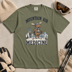 Flat lay of a moss green t-shirt featuring a peaceful moose ski graphic with “Mountain Air Medicine” text, styled with winter accessories.