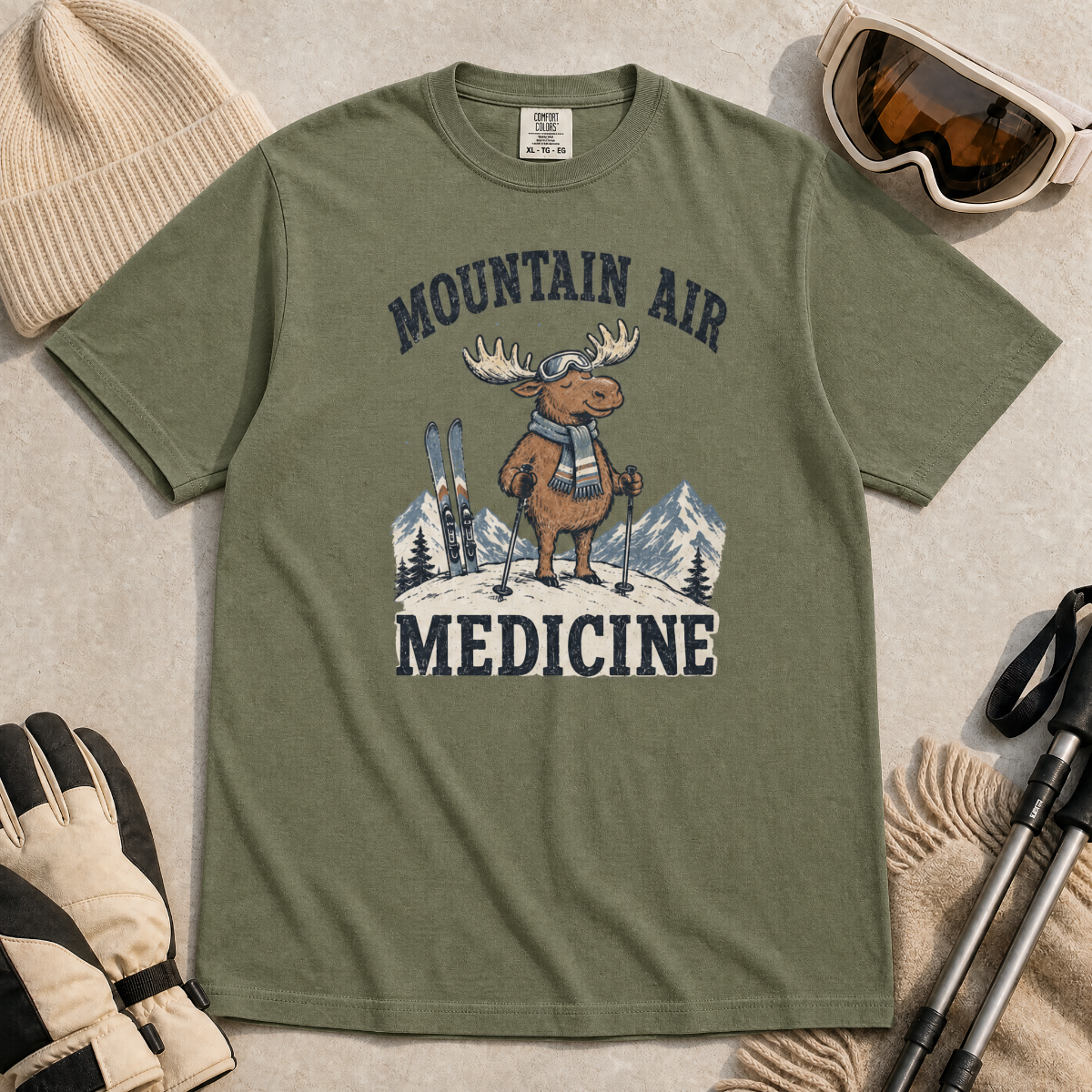 Flat lay of a moss green t-shirt featuring a peaceful moose ski graphic with “Mountain Air Medicine” text, styled with winter accessories.