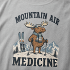 Close-up of a granite gray t-shirt with a retro ski design showing a calm moose with goggles, scarf, ski poles, and mountain scenery, with “Mountain Air Medicine” text.
