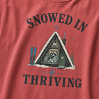 Close-up of a crimson red t-shirt with a vintage ski cabin design showing a cozy raccoon wrapped in a blanket inside an A-frame cabin, with “Snowed In & Thriving” text.
