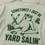 Close-up of a bay green t-shirt with a green vintage ski graphic showing a worn-out bear after a yard sale crash, surrounded by skis, goggles, poles, and a boot.