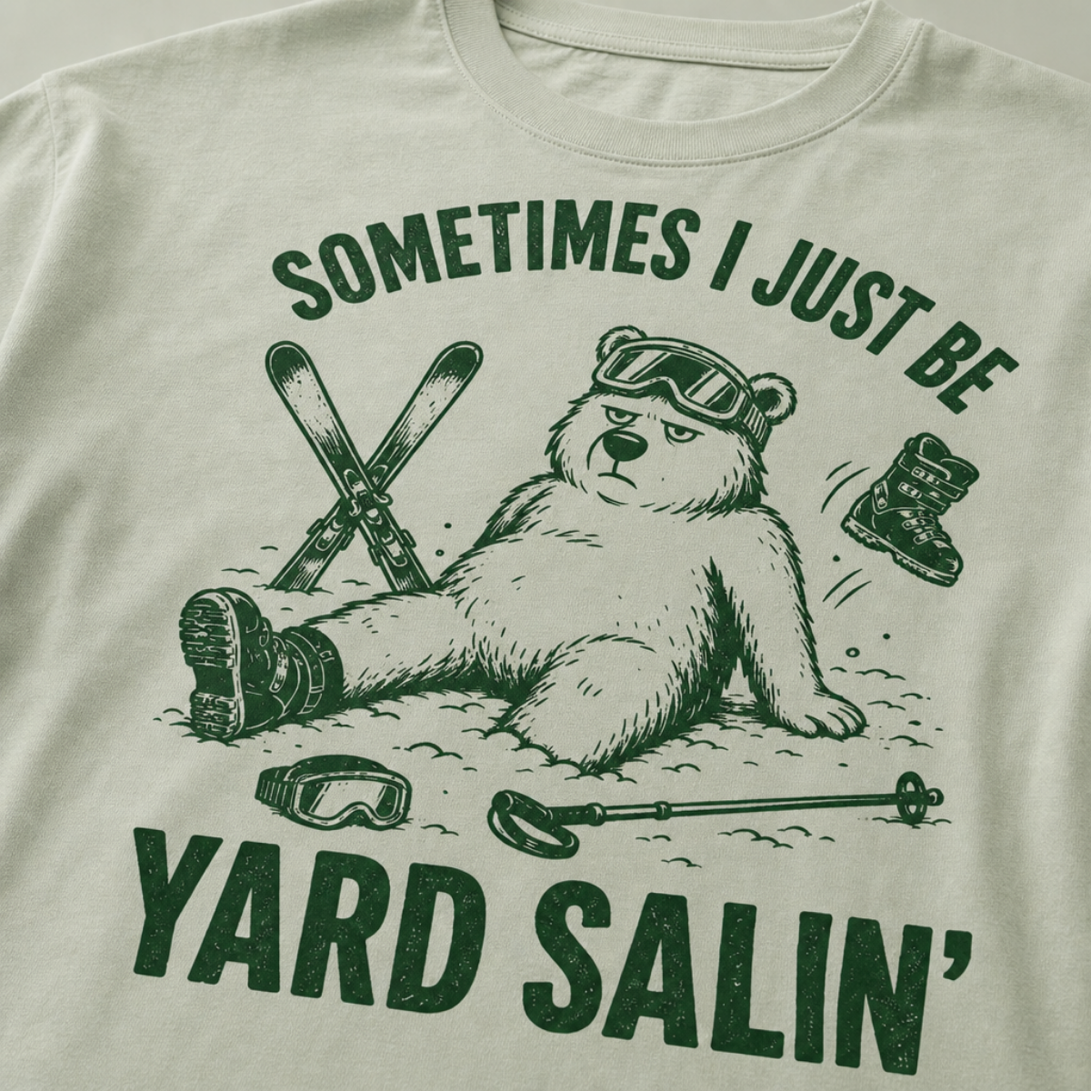 Close-up of a bay green t-shirt with a green vintage ski graphic showing a worn-out bear after a yard sale crash, surrounded by skis, goggles, poles, and a boot.
