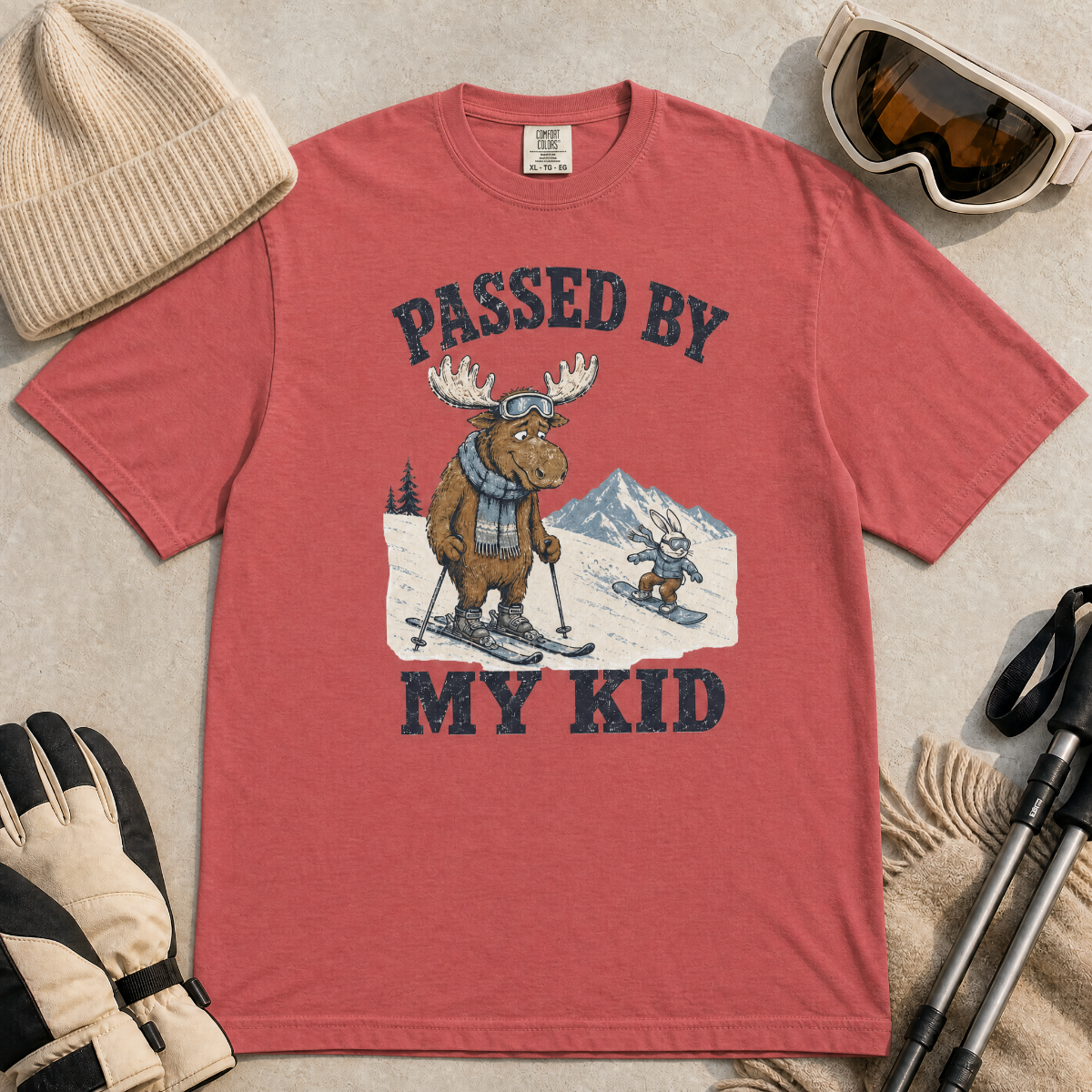 Flat lay of a crimson red t-shirt featuring a funny “Passed By My Kid” ski design with a humbled moose watching a small rabbit snowboarder pass by.