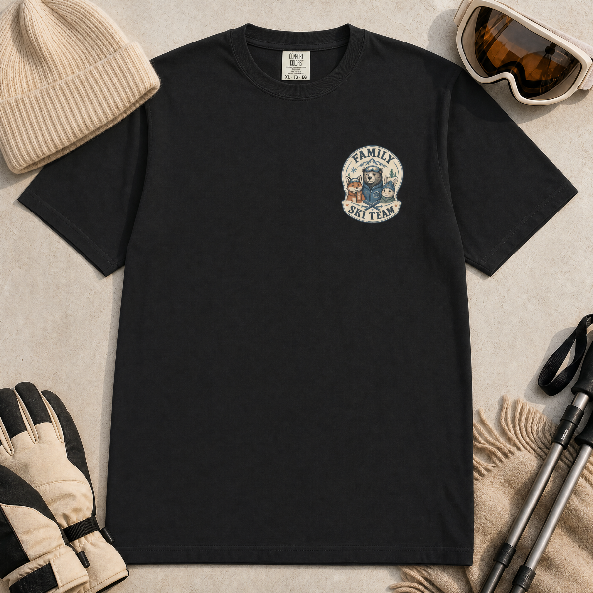 Flat lay of a black t-shirt featuring a small chest “Family Ski Team” badge graphic with three winter animal characters, styled with ski accessories.