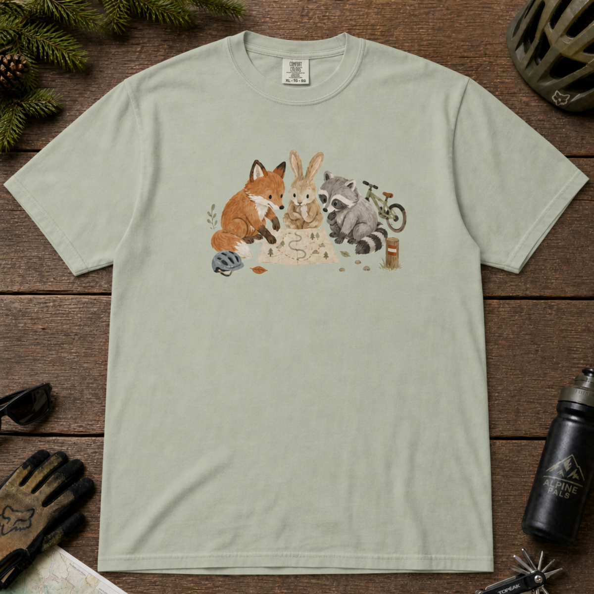 Flat lay of a bay green t-shirt featuring a cute woodland animal trail-planning graphic with a fox, rabbit, raccoon, map, helmet, bike, and trail marker.