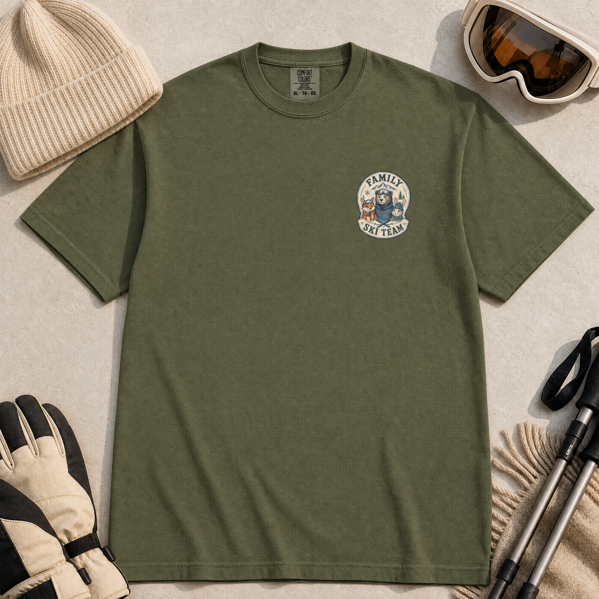 Flat lay of a hemp green t-shirt featuring a small chest “Family Ski Team” badge graphic with three winter animal characters, styled with ski accessories.