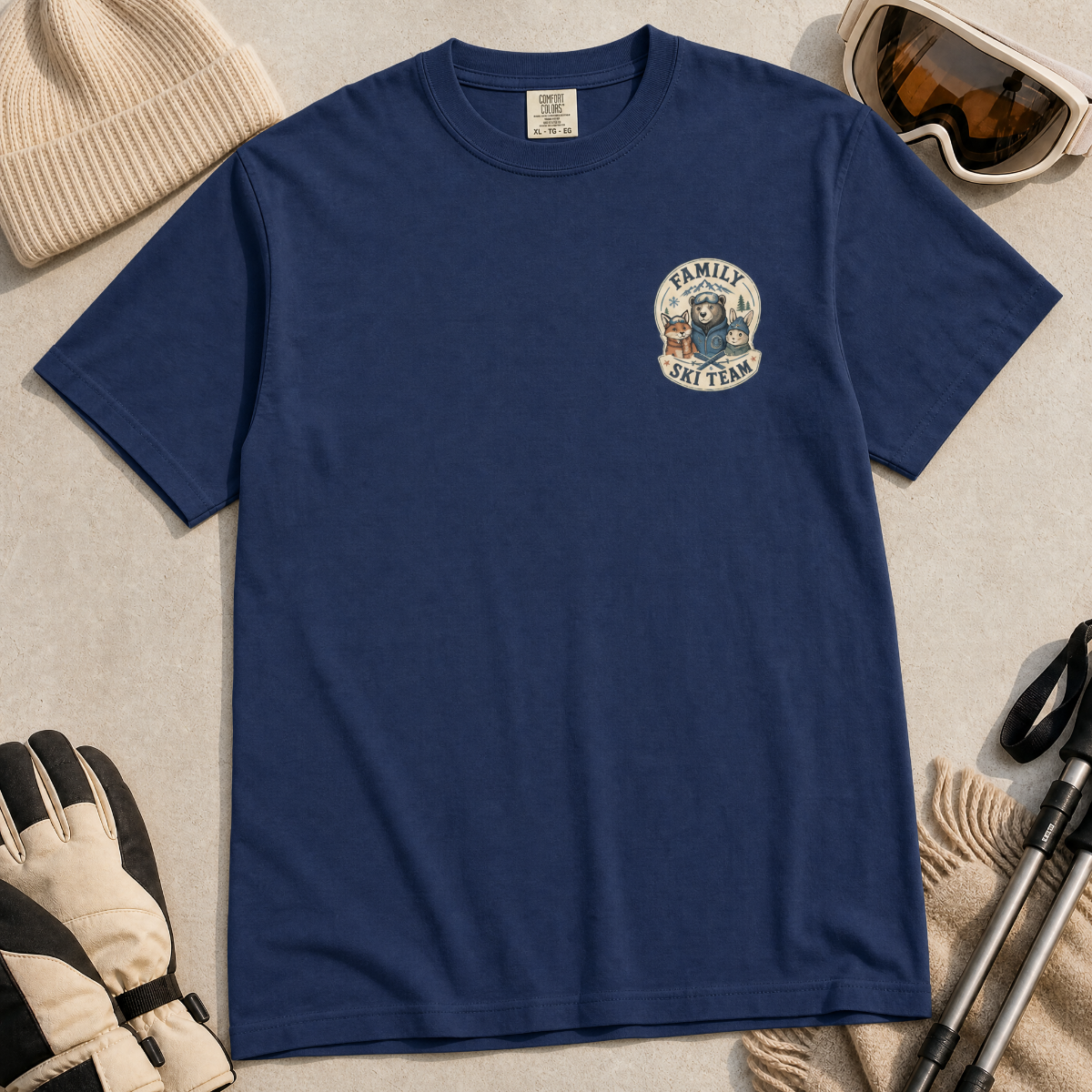Flat lay of a navy blue t-shirt featuring a small chest “Family Ski Team” badge graphic with three winter animal characters, styled with ski accessories.