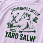 Close-up of a orchid colored t-shirt with a green vintage ski graphic showing a worn-out bear after a yard sale crash, surrounded by skis, goggles, poles, and a boot.
