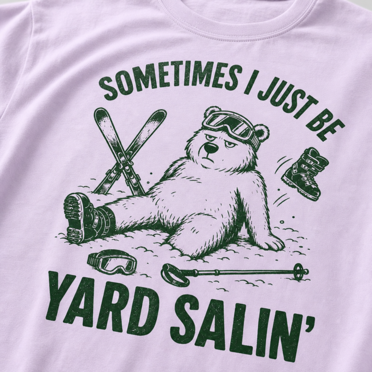 Close-up of a orchid colored t-shirt with a green vintage ski graphic showing a worn-out bear after a yard sale crash, surrounded by skis, goggles, poles, and a boot.