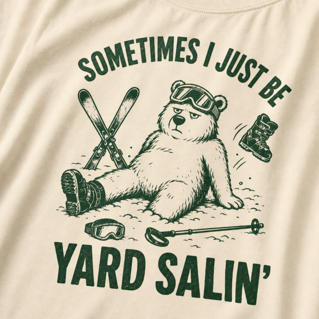 Close-up of an ivory white t-shirt with a green vintage ski graphic showing a worn-out bear after a yard sale crash, surrounded by skis, goggles, poles, and a boot.