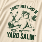 Close-up of an ivory white t-shirt with a green vintage ski graphic showing a worn-out bear after a yard sale crash, surrounded by skis, goggles, poles, and a boot.